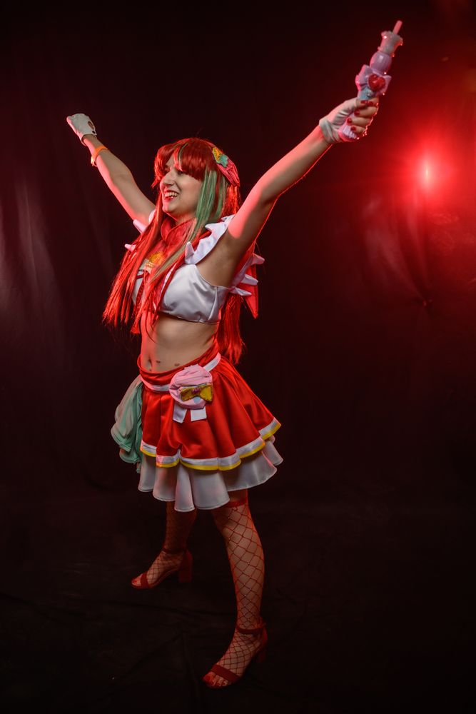 Fanelia is pictured cosplaying as Cure Flamingo from Tropical Rouge Precure. Cure Flamingo is in her “Victory!” pose. The photo was taken by Huffoto!