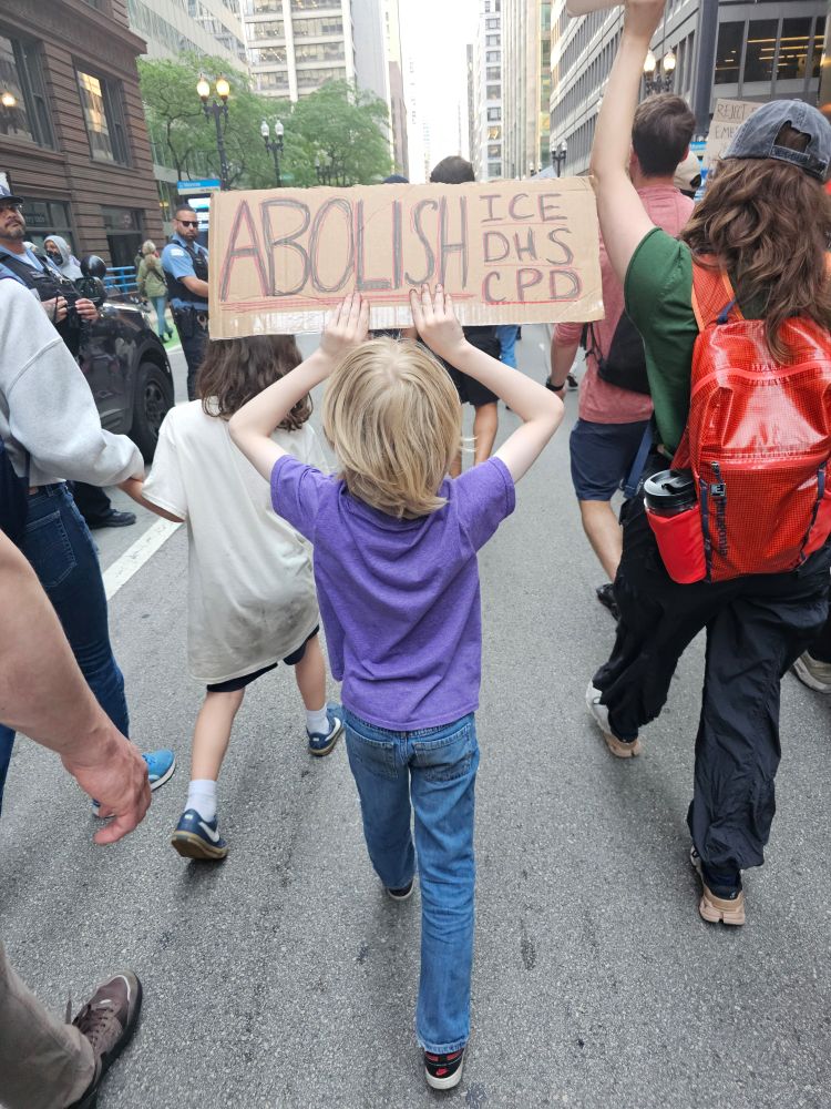 An 8-year-old boy with blonde, chin length hair, a purple shirt and blue jeans marches in the Chicago ICE OUT protest. He holds a sign that says ABOLISH ICE, DHS, CPD. 