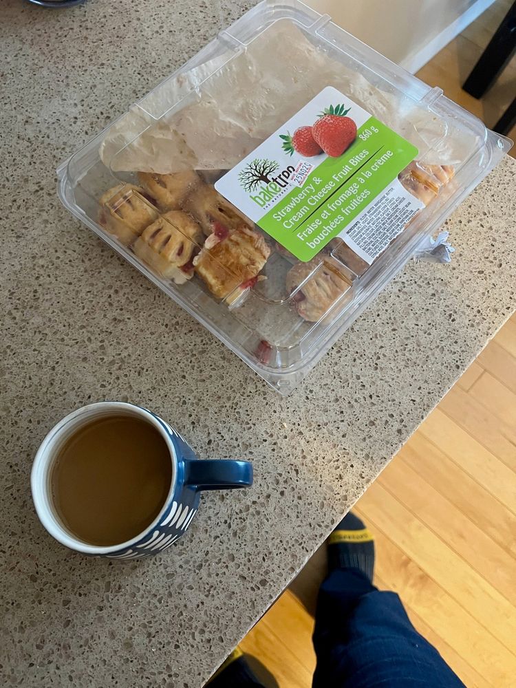 A cup of coffee and an open container of strawberry and fruit bars are placed on a kitchen counter with someone's feet visible in the corner.