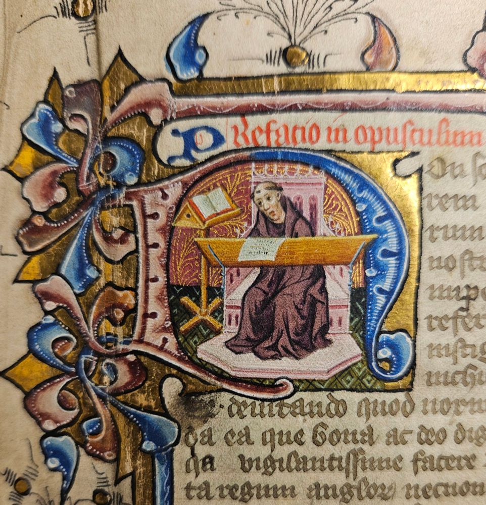 An illuminated letter N inside which is a monk, writing, with a mildly bemused look on his face.