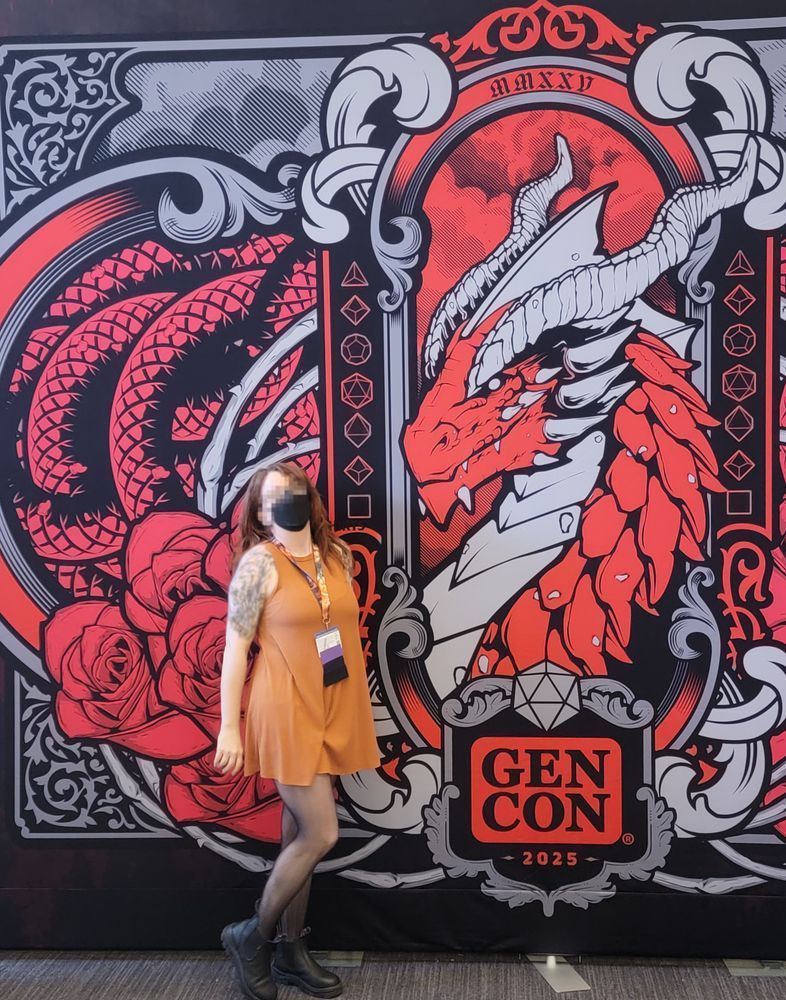 A pale, tattooed redhead wearing a black kf95 mask, short orange dress, fishnets and black boots standing in front of the large red dragon art for GenCon 2025