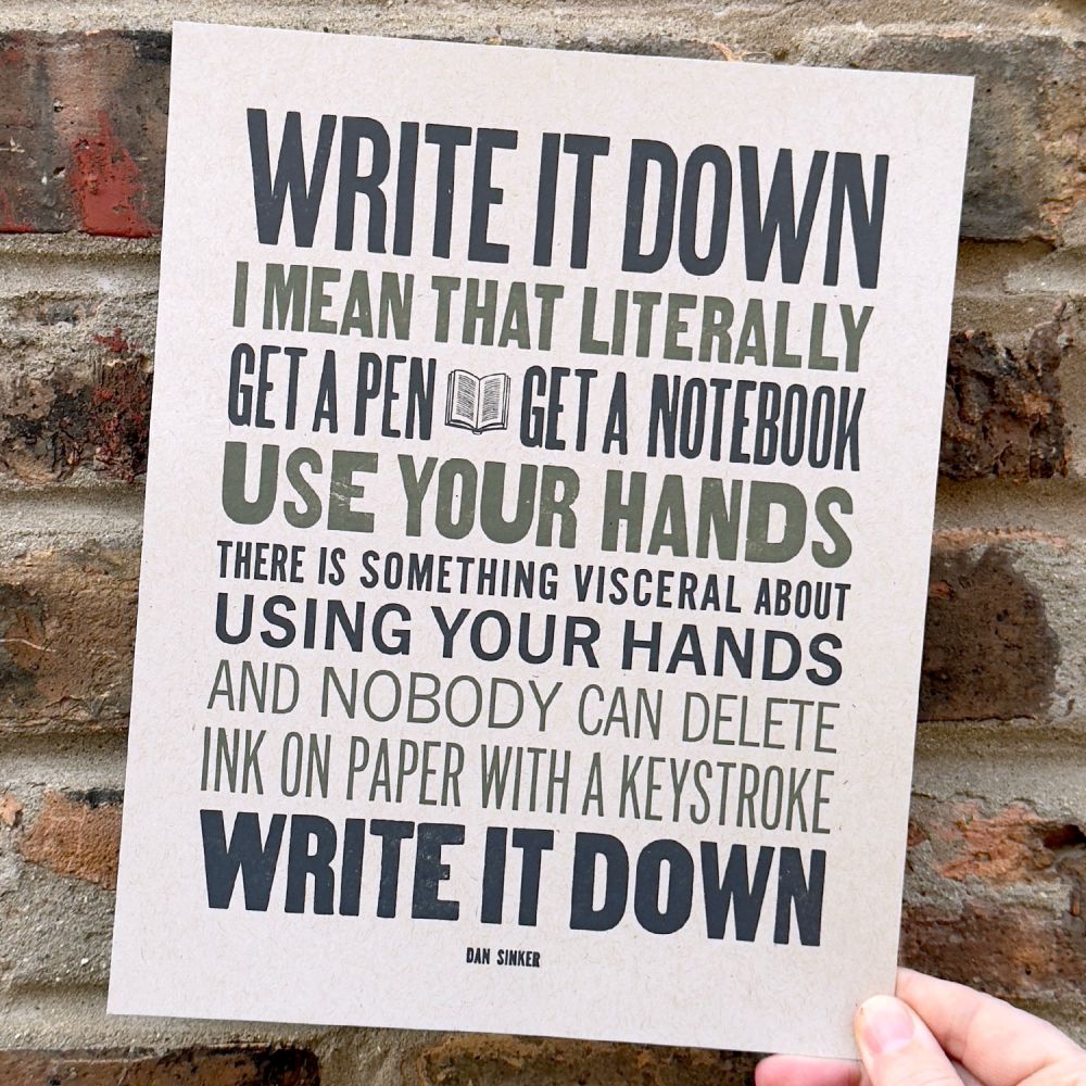 A hand holding an 8x10 print with a quote from Dan Sinker that says ‘write it down. I mean that literally. Get a pen, get a notebook, use your hands. There’s something visceral about using your hands. And nobody can delete ink on paper with a keystroke. Write it down.’