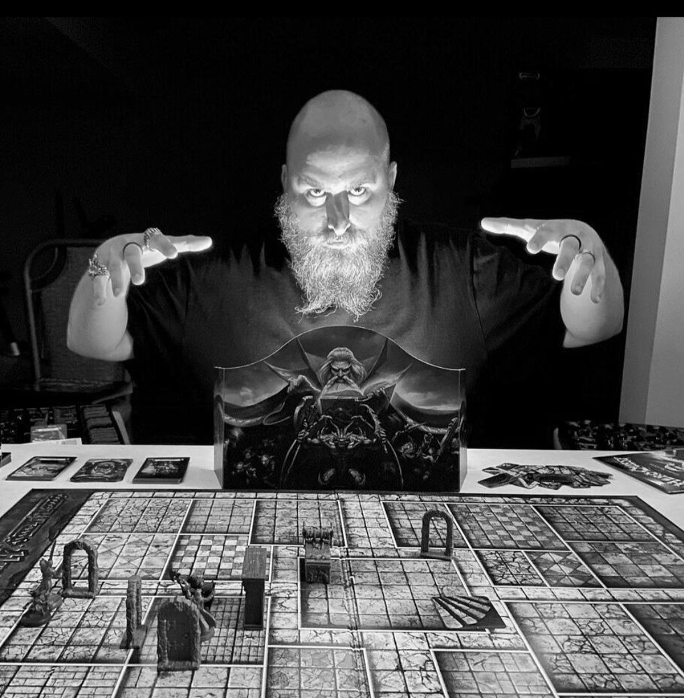 A bald man with a beard is sitting at a table, his hands extended over a game of Heroquest