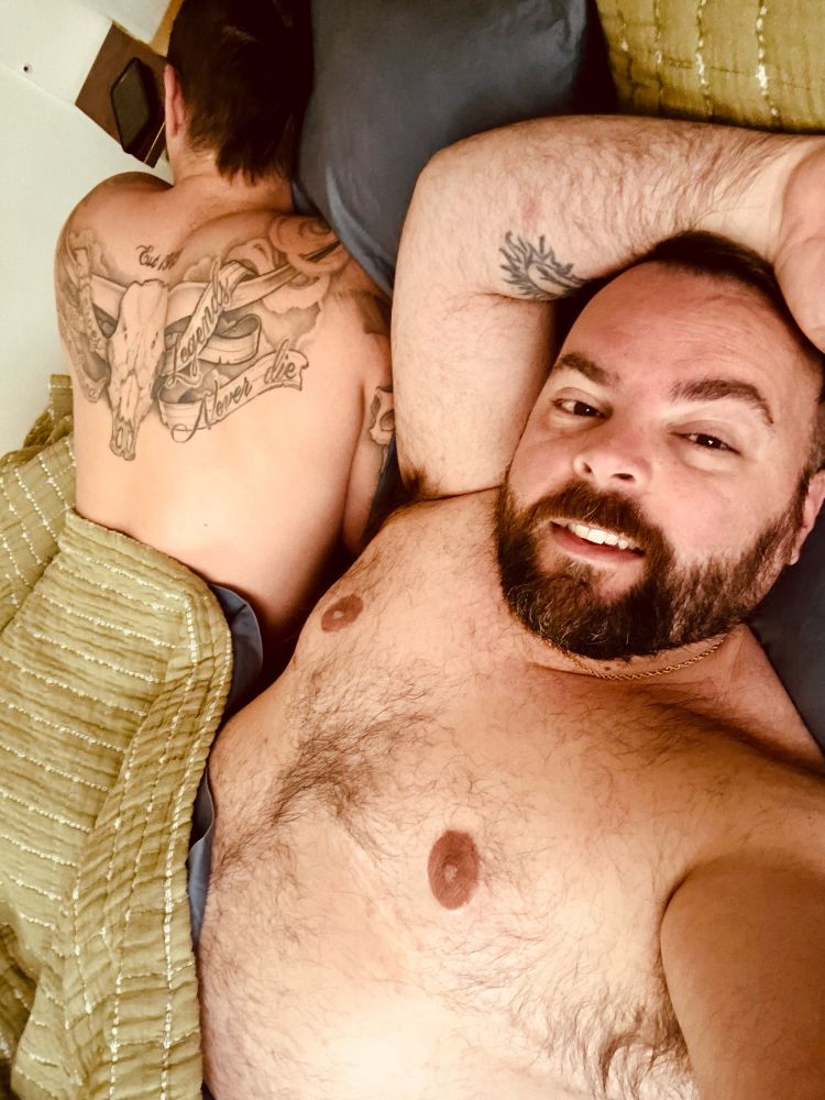 Two men in bed, one face toward camera the other away sleeping 
