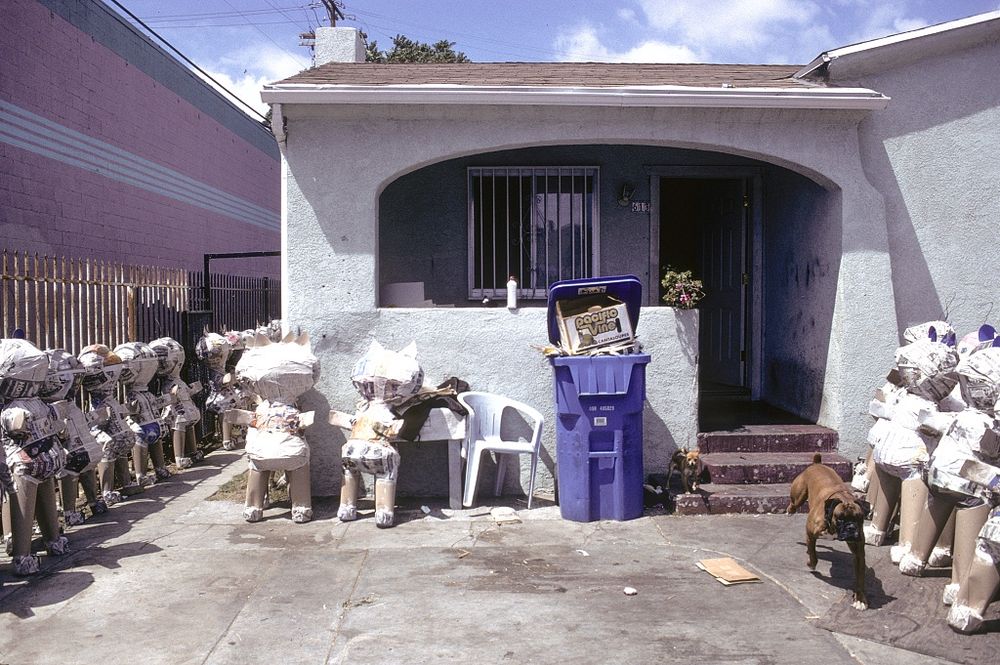 Before an LA bungalow stand multiple piñatas in progress. To one side, a dog appears to run toward the photographer.