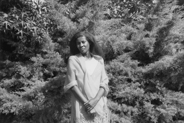 A vintage black and white image shows Alice Coltrane with her hands clasped, standing before a forested landscape.