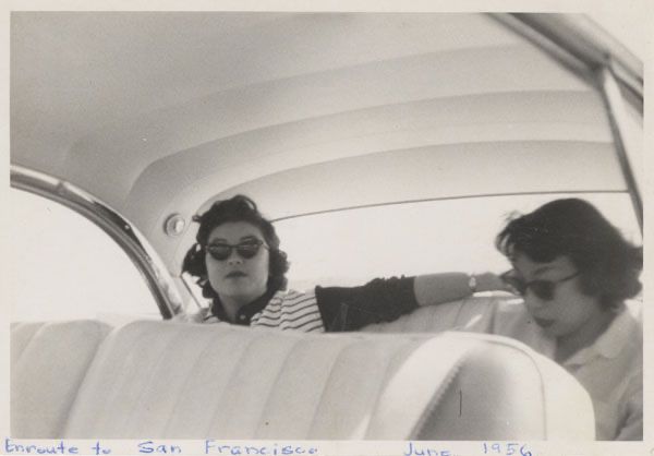 A vintage black and white snapshot shows two Japanese American women in cat eye sunglasses sitting inside a beautifully upholstered Chevy Bel Air.