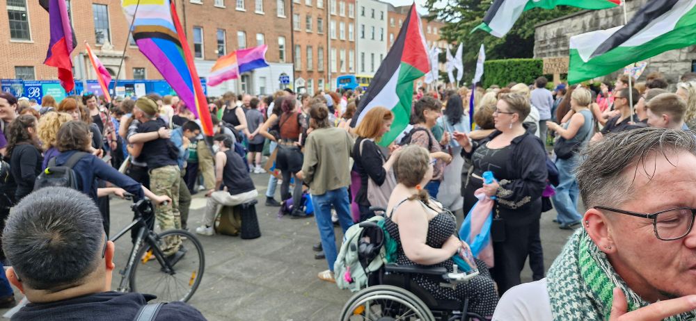 Happy dykes & supporters fill the space outside the gates of the Garden of Remembrance. There are many queer flags, but many Palestinian flags also.