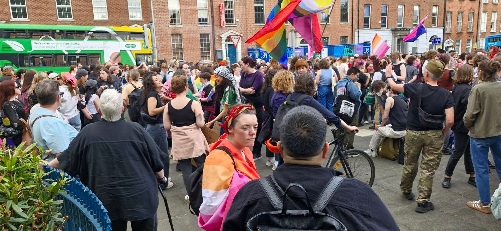 Happy dykes & supporters fill the space outside the gates of the Garden of Remembrance. There are many queer flags, but many Palestinian flags also.