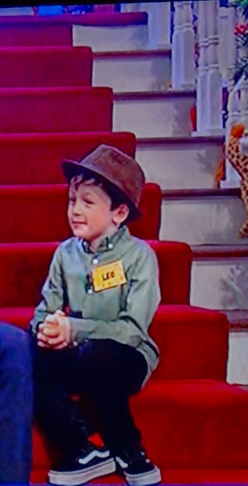 Leo, a young storyteller from Leitrim, sits on the steps of the Late Late Toy Show set, wearing a hat. 