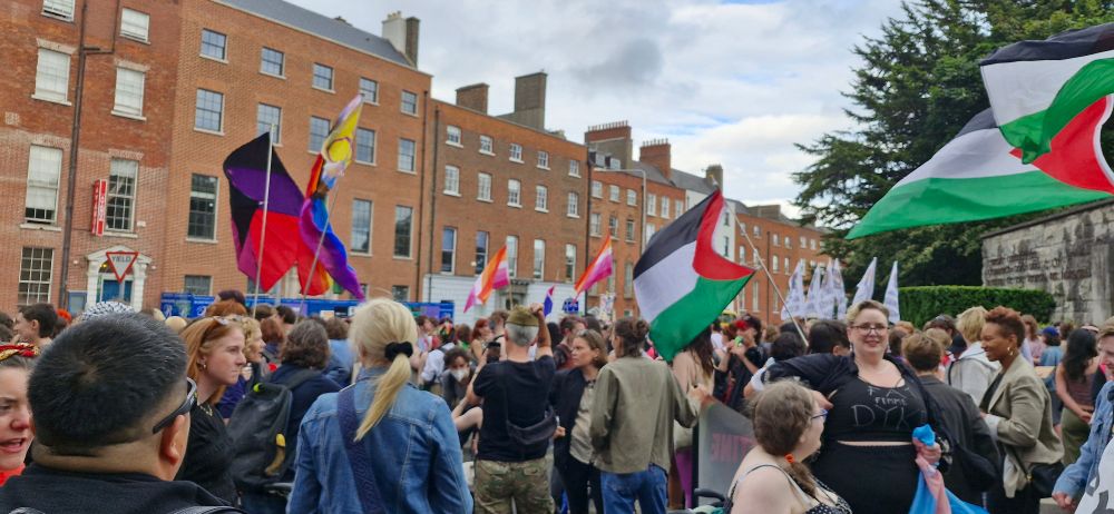 Happy dykes & supporters fill the space outside the gates of the Garden of Remembrance. There are many queer flags, but many Palestinian flags also. 