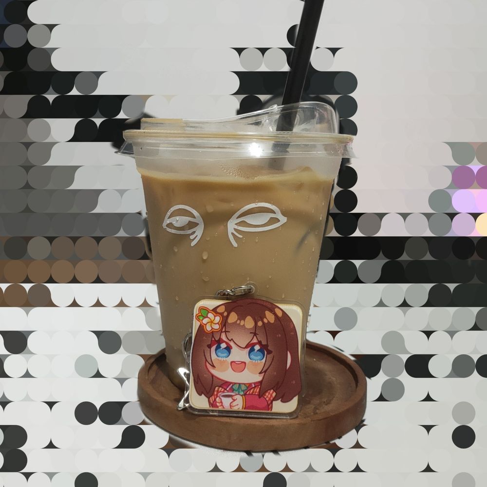 A picture of a cup of iced latte and a Hana Macchia (Nijisanji ID) keychain in front of it