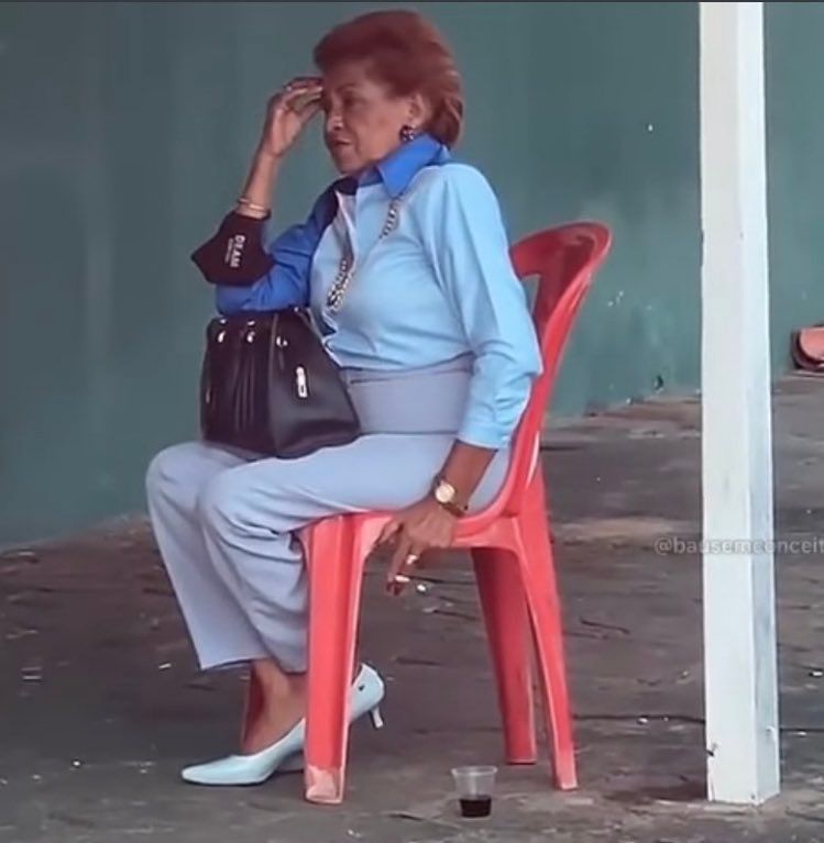 uma senhora muito elegante em uma roupa social azul com sapato de salto branco e uma bolsa preta, está sentada em uma cadeira vermelha de bar fumando um cigarro e tomando um copinho de café preto 