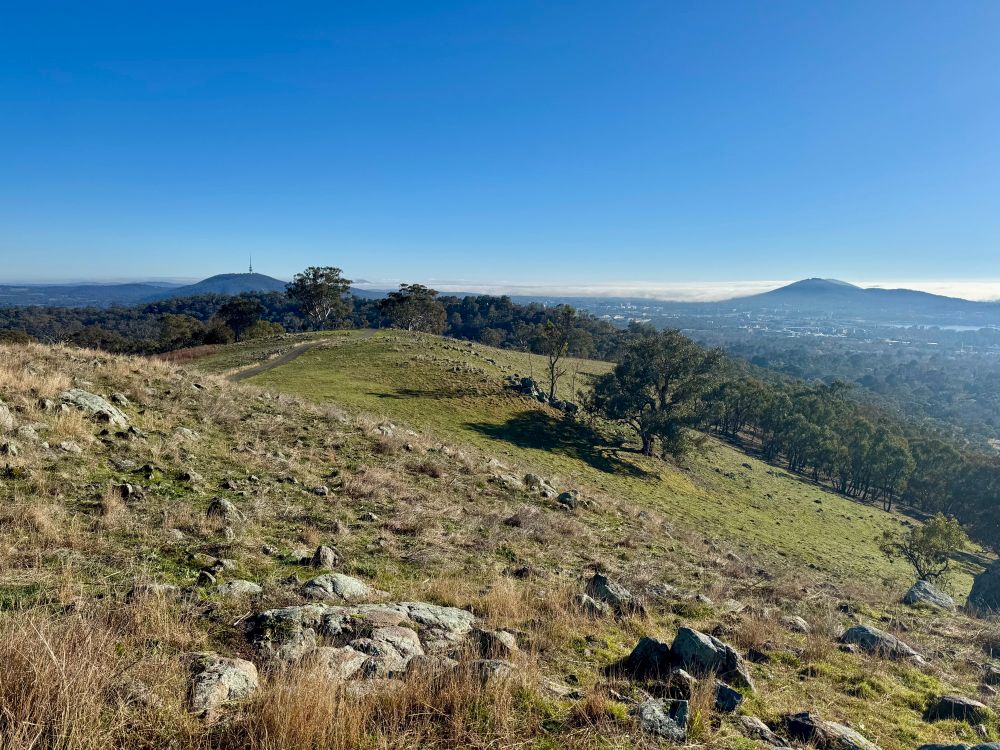 View from the trig point atop Red Hill in Canberra. The sky is blue. There’s a light covering of grass on the hills and a thin layer of cloud in the distance. You can just see the Black Mountain tower off to the left. There are rocks. 