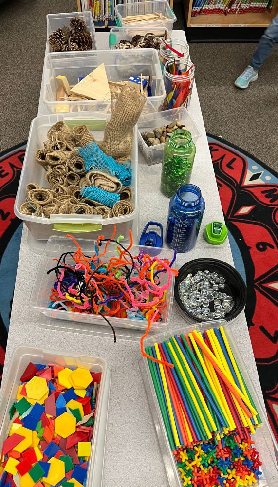 A bunch of loose parts building items laid out on a table in their separate bins: plastic shapes blocks, straws and connecters, pipe cleaners, gems that are clear or green or blue, little rolls of burlap, twigs and wooden sticks, etc…