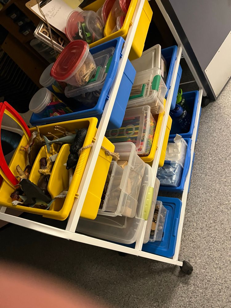 The “loose parts” cart.
It has rolling wheels for easy transporting, three tiers of shelves to stack bins on, and is approximately 1 foot wide by 4 feet long.
Mostly, the goal is to have non-representational objects (other than the plastic animals and some toy cars, which often aren’t needed/used) to build with imaginative creativity.
Bins of paper tubes, various building block items, pipe cleaners, feathers, burlap strips, ribbons, pine cones, various sticks items…etc, etc.
