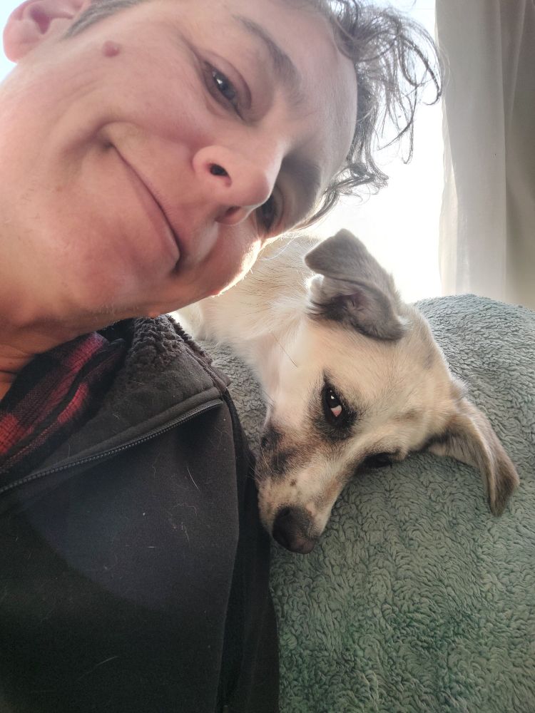 Charlie is leaning his head over to his right where Delia the dog is resting over the back of the chair. Charlie is smiling and Delia looks like she's giving him side eye