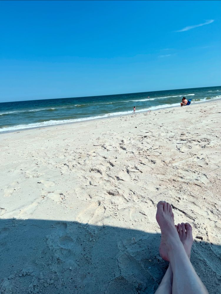 A beach scene. Sand stretching to the ocean. Gentle rolling waves with whitecaps. Other beachgoers in the distance. 