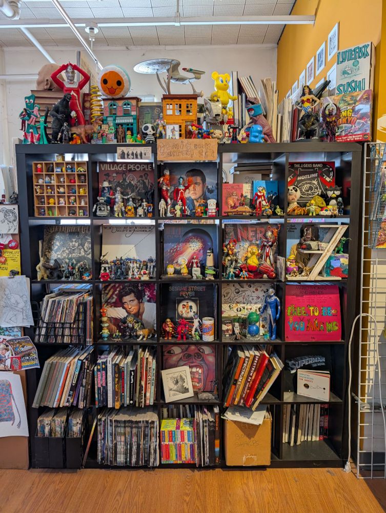 A grid-like display of comics, toys, records, and various other paraphernalia, on an Ikea shelf inside World Monster HQ. 