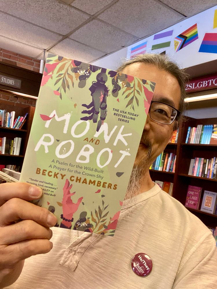 Picture of a bookseller in glasses in a beige top holding up a copy of “Monk and Robot” by Becky Chambers (ISBN 9781250386335), a trade paperback omnibus edition that includes “A Psalm for the Wild-Built” and “A Prayer for the Crown-Shy” featuring a light green cover with the title in white handwriting-style lettering and a human hand reaching up from below and a grey robot hand reaching down from above. 