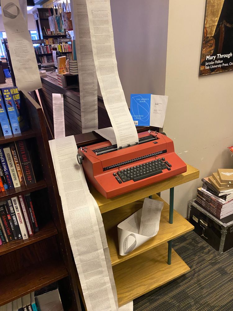 Pictured is the opposite side of the stairwell opening in the first photo, showing a reddish orange vintage typewriter on a stand out of which is flowing a scroll of paper. A later section of the scroll can be seen flowing back over the edge of the opening beside the typewriter. There is also a smaller Möbius strip, made up of the front and back matter of the book, sitting on a shelf below the typewriter. 