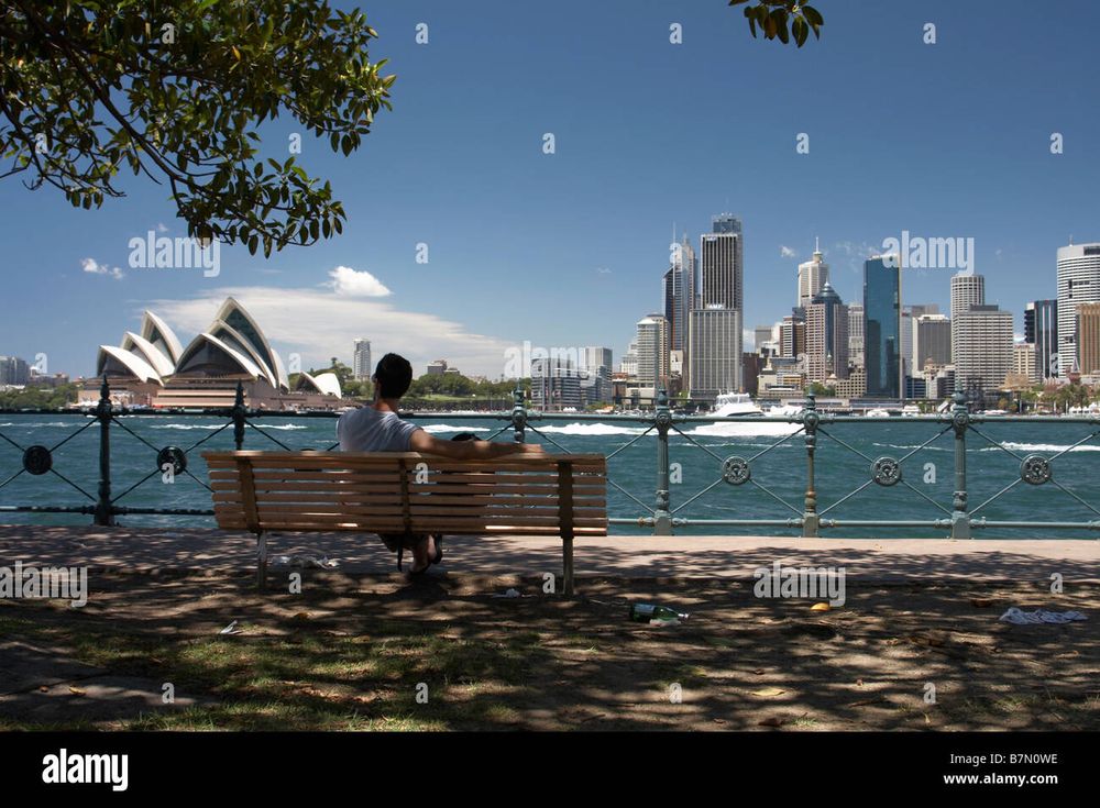 View from Kirribilli on north side of Sydney harbor towards skyscrapers and Sydney Opera House across the water. A man sits on bench surveying the view