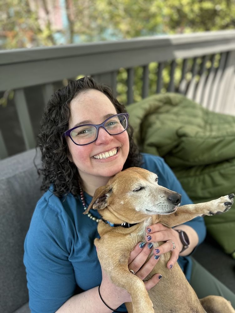 Happy white woman with dark curly hair is sitting on an outdoor couch, holding up a sleepy dachshund-mix. 
