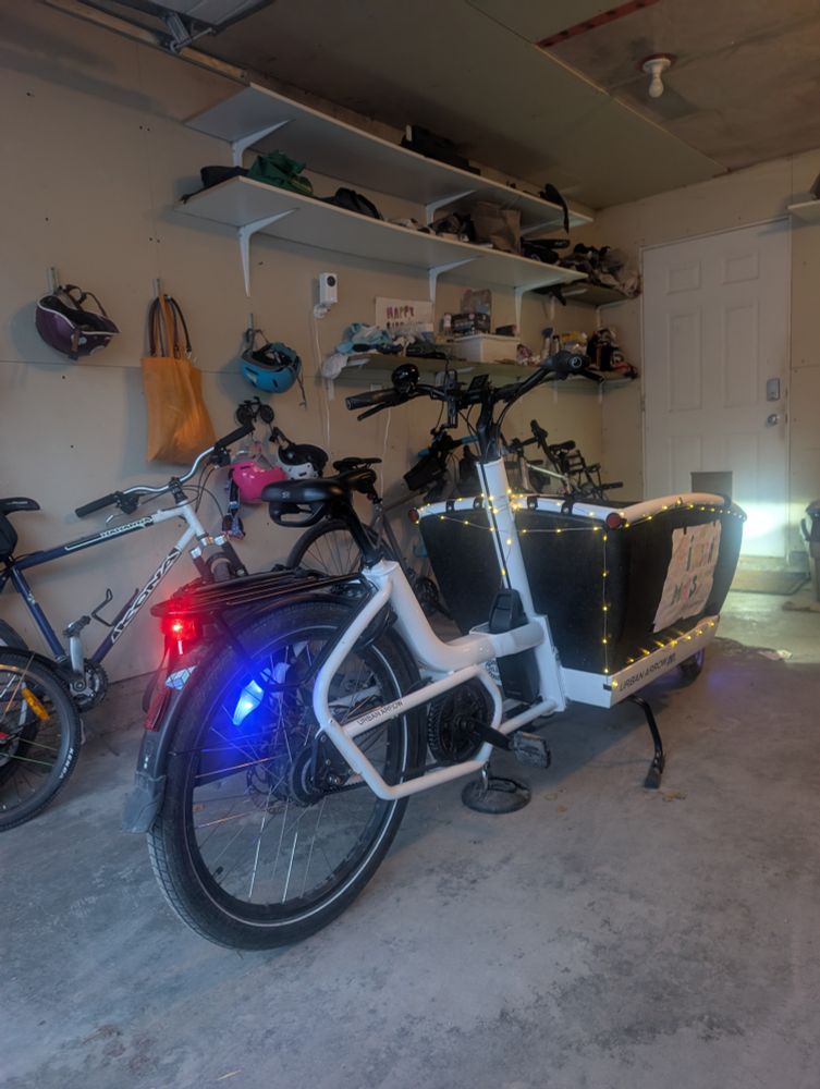 An Urban Arrow cargo bike parked in a garage with Christmas lights around the box and spoke lights on the wheels. 