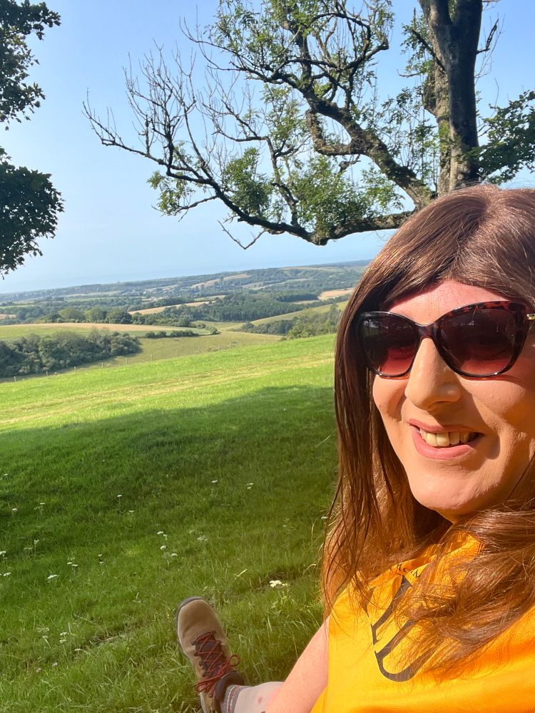 Emily with the south coast behind her sitting in chanctonbury ring, a ‘haunted’ Neolithic hill fort