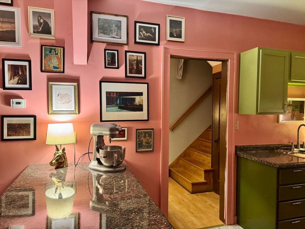 a pink wall covered in art, dark green lower cabinets, and lighter green upper cabinets. there is a doorway in the middle of the wall looking onto some stairs