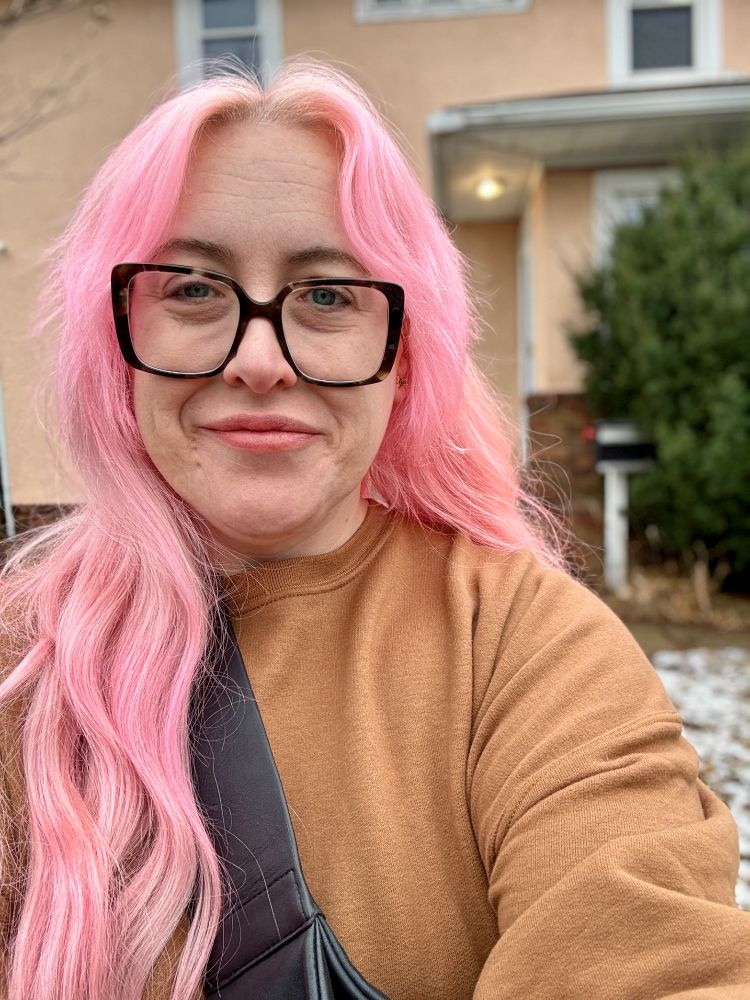 a pink-haired woman in a brown sweatshirt standing in front of a pink house
