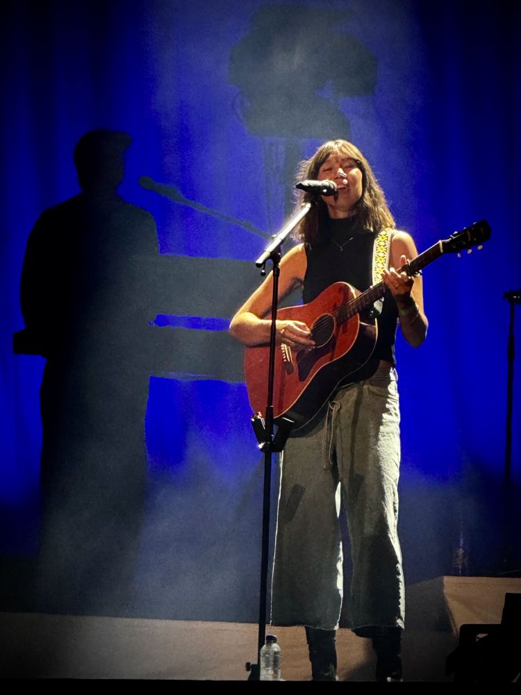 Zangeres Beau zingt en speelt gitaar op een podium met blauw licht.