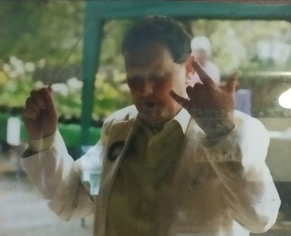 Our Da, who had dark hair, wearing a lemon shirt, a cravat, & a pale linen suit with a local safety campaign sticker on the lapel, holding a baton. Pictured while conducting the Erme Singers at an open air concert in aid of the survivors of the Tsunami.