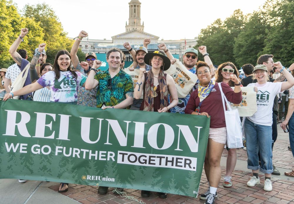 A group of people wearing REI union gear and fun union clothing holding a banner that says: REI Union - We Go Further Together. 