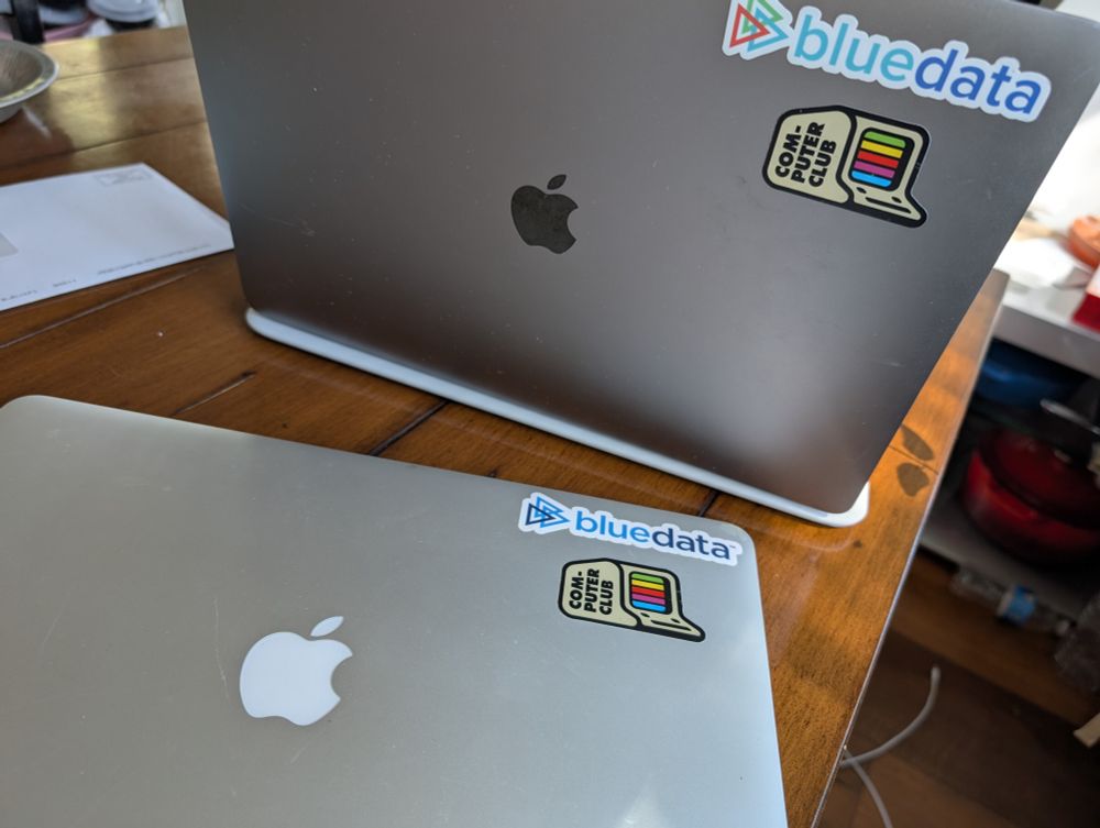 Two old macbooks. The cover of each is sporting a sticker in the shape of an old personal computer. That computer image is labelled with the text "COMPUTER CLUB", and it shows a chunky rainbow on its screen similar to the colors of the old Apple logo.