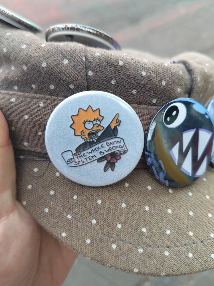 A grey hat with a pin of Lisa from The Simpsons saying "THE WHOLE DAMN SYSTEM IS WRONG!".