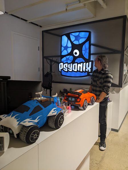 Donny, wearing a grey flannel and black pants, turning to look at the bright blue Psyonix logo behind them. In front of them is a shelf with two giant stuffed rocket league cars on it