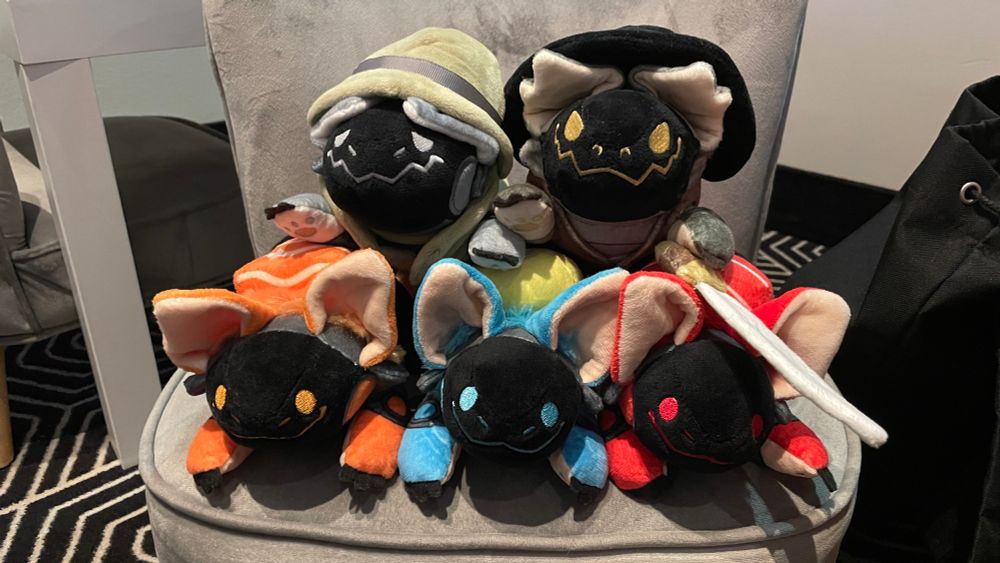 5 protobean plushies stacked as a pyramid on a child’s chair, the plushies have some costumes like one i a solder, another like crocodile dundee and three are different types of sushi 