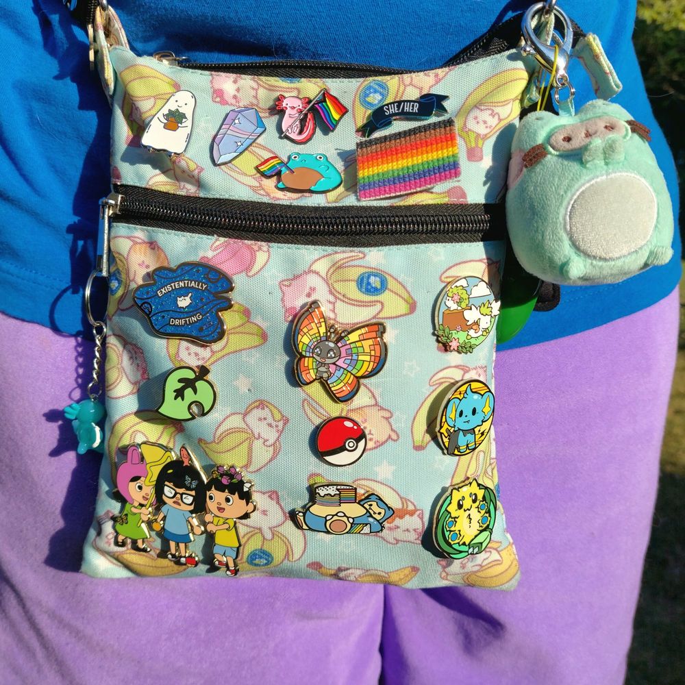 Photo of a small crossbody bag with a Bananya design (cute cartoon cats in banana peels) and covered with mostly enamel pins. The pin designs include Pokémon, Bob's Burgers, Animal Crossing, LGBTQ+, and other themes. There is a pronoun pin that says "she/her" and a galaxy pin with a floating cat that says "existentially drifting." Cute keychains also hang from the purse straps and zippers. The person, me, wearing the purse is wearing a blue shirt and purple shorts and is standing outside on a sunny day.