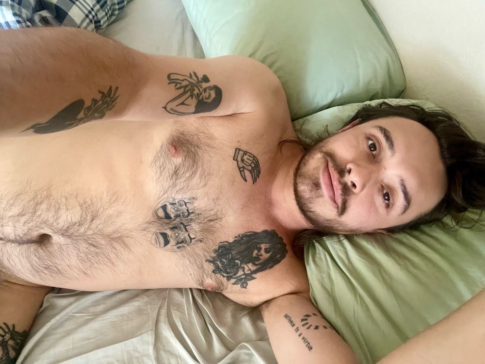 Me laying down in bed without any clothes in the morning as I’m waking up. My arms and chest are covered in tattoos, and I am laying on light green pillows. 