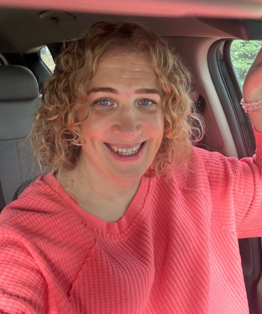 Woman with curly blonde hair sitting in a car. She is wearing an oversized salmon color top and is smiling. 