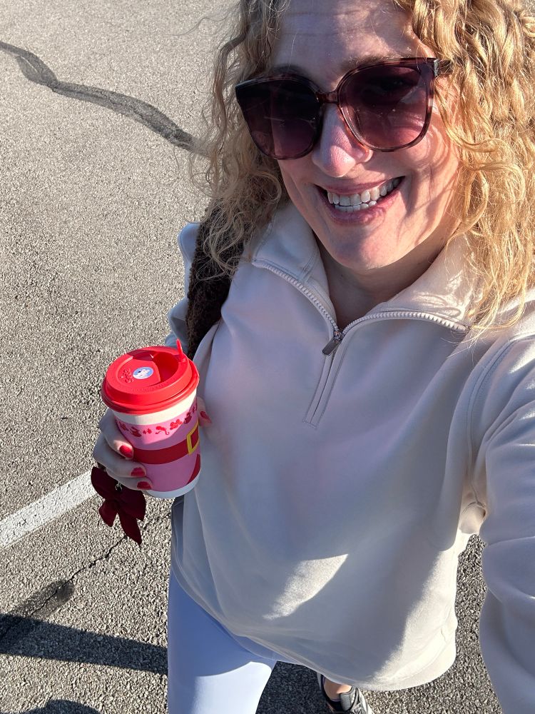 Woman in light blue yoga pants and a cream pullover. She’s holding a coffee and wearing sunglasses. 