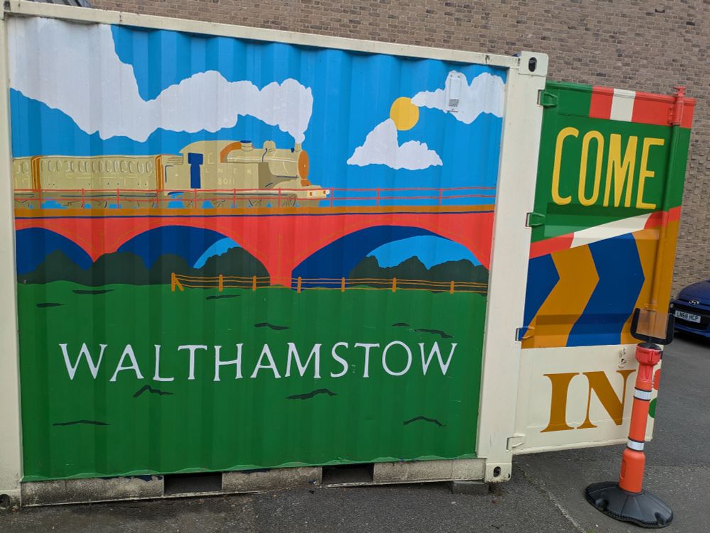 A shipping container painted with a depiction of a train passing over a viaduct, the fields below say "Walthamstow", the open doors say "Come In"