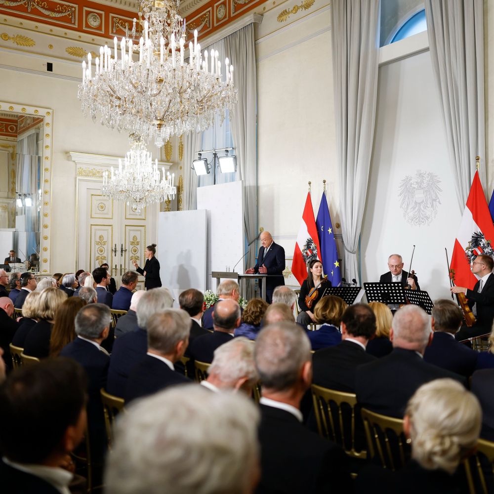 Saal mit Publikum, Bundeskanzler Christian Stocker hält eine Rede, rechts sitzen Musiker:innen mit Instrumenten. 