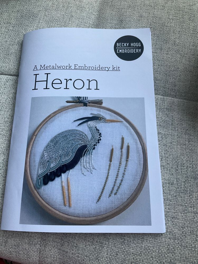 A booklet for a Metalwork Embroidery kit by Becky Hogg. The photo on the front shows a heron stitched mainly in silver on a white fabric background in a small embroidery hoop. The heron has dark blue and teal accents and it is standing next to three bulrushes.