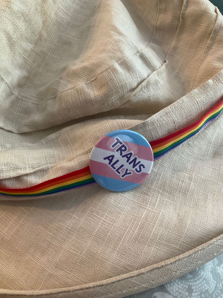 A beige fabric hat with a pride ribbon around the crown and a trans ally badge pinned to it.