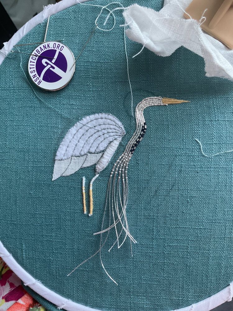 A metal thread embroidery (Goldwork but not gold) of a heron in progress on a teal fabric. The felt and soft string padding can be seen and the beak and legs are complete. The head and neck are being couched using silver thread sewn down with white thread with blue and teal making a pattern on the neck. There is a RSN Stitchbank magnetic needle keeper to the top left.