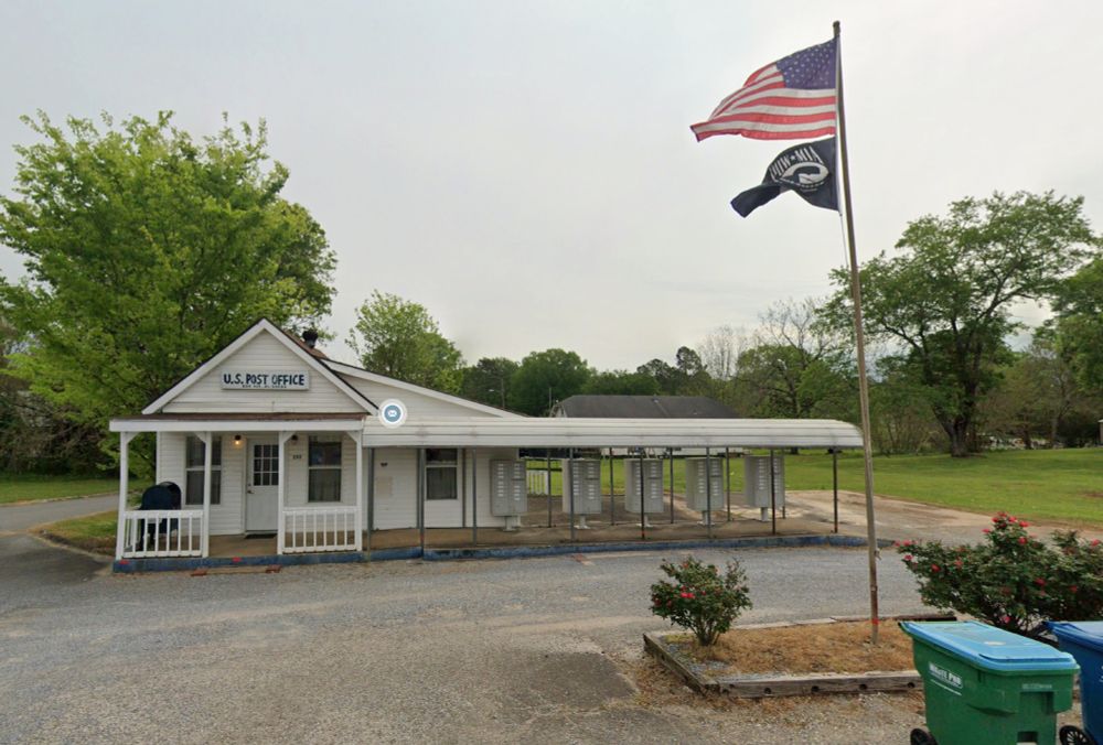 A small, one-story white building with a hand-painted blue sign reading, "U.S. Post Office, Bon Air, AL" with a small gravel parking lot in front. There are five sets of cluster mailboxes under a roof to the right, and U.S. and POW-MIA flags are flying overhead.