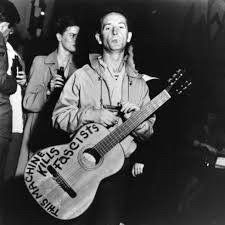 Black and white photo of Woodie Guthrie with acoustic with the words painted on it “this machine kills fascists”
