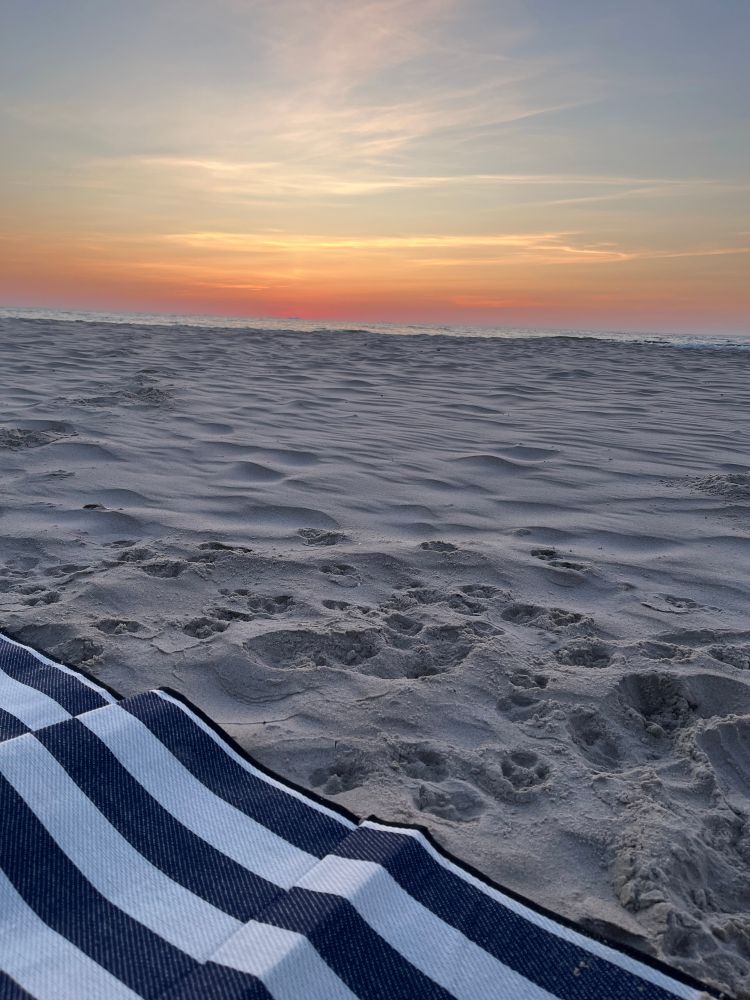 Ein Stück blau-weiß gestreifte Decke an einem einsamen Strand. Fuß- und Pfotenspuren im Sand, über dem Meer ist der Himmel Rotgold. Die Sonne geht bald auf